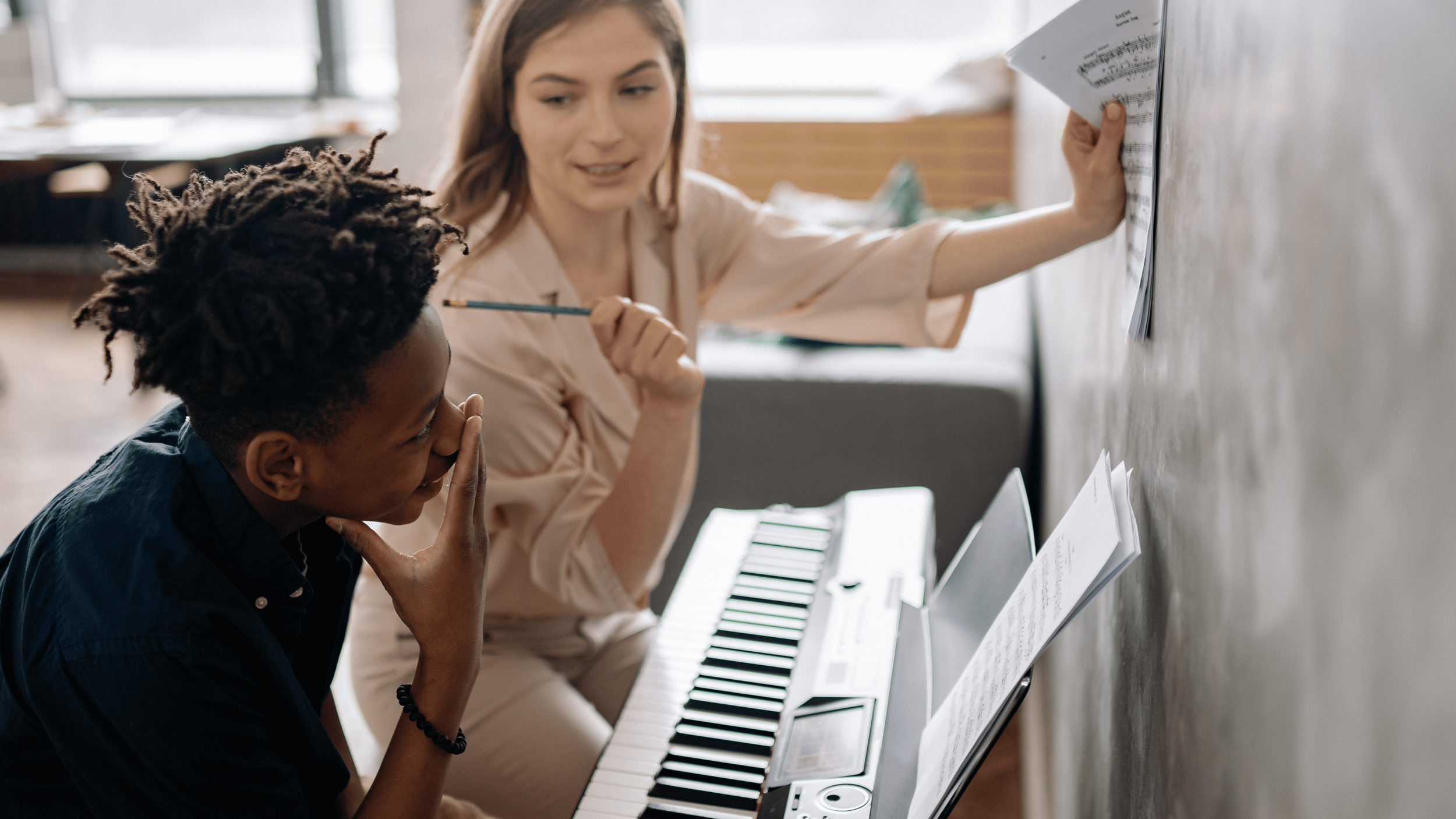 Teacher pointing at sheet music on a wall while a student sits at a keyboard, listening and thinking during a lesson.