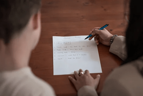 Person writing notes on a paper titled “Music Ideas” while a child looks on across a wooden table.