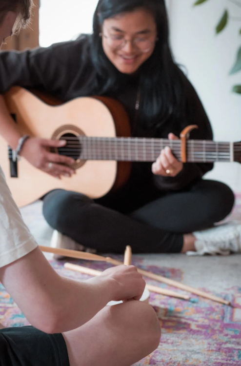 Person playing an acoustic guitar while a child taps drumsticks on a small drum during a music session.