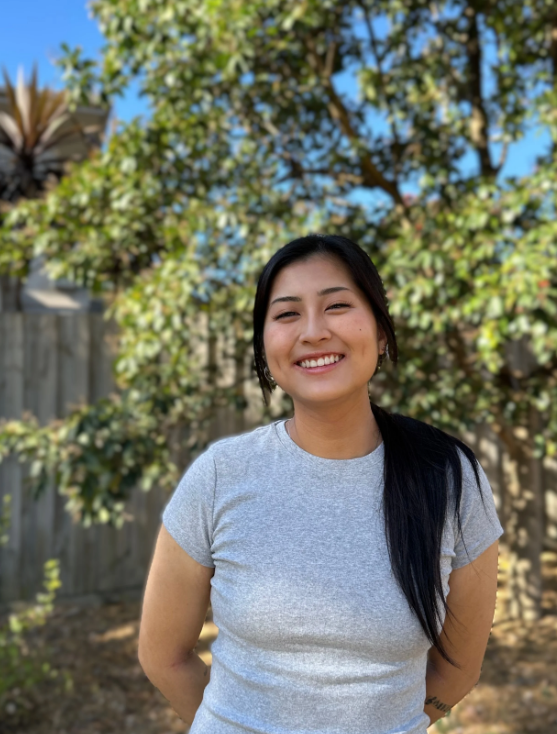 Smiling person standing outdoors in a sunny yard with trees and a wooden fence in the background.