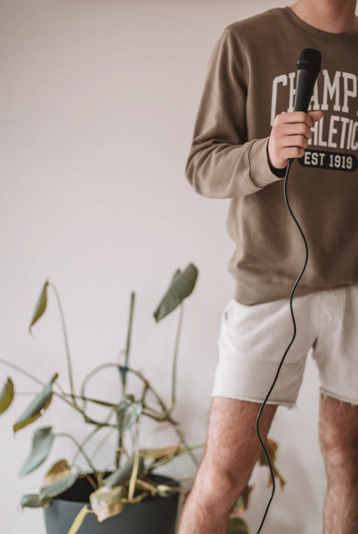 Person holding a wired microphone, wearing a brown sweatshirt and white shorts, standing indoors beside a potted plant.