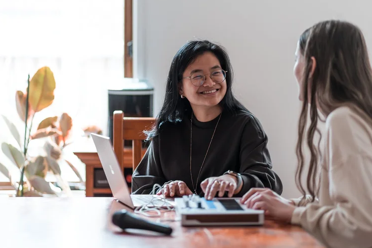Two people smile and collaborate at a table using a laptop and small music controller in a bright room.