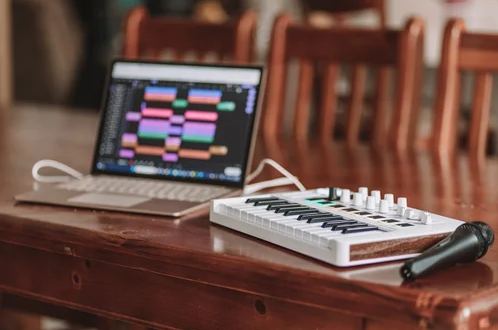 Laptop displaying music production software beside a small MIDI keyboard controller and microphone on a wooden table.