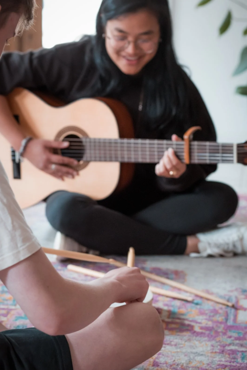 Person sitting cross-legged playing an acoustic guitar while a child taps drumsticks on a small drum during a music activity.
