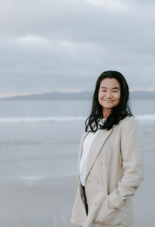 Person in a light coat smiling at the camera while standing on a cloudy beach with ocean waves in the background.