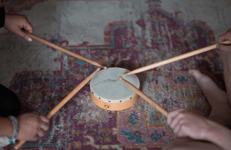 Four people tap drumsticks on a small tabletop drum placed on a patterned rug during a group activity.