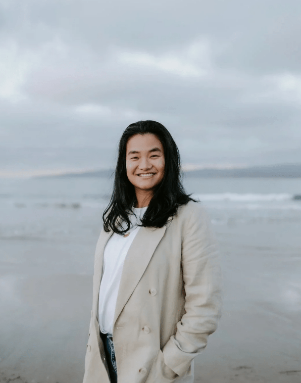 Person in a light coat smiling at the camera while standing on a cloudy beach with ocean waves behind.