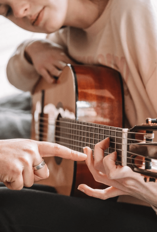 Instructor pointing at a child’s fingers on an acoustic guitar fretboard while teaching how to form a chord.