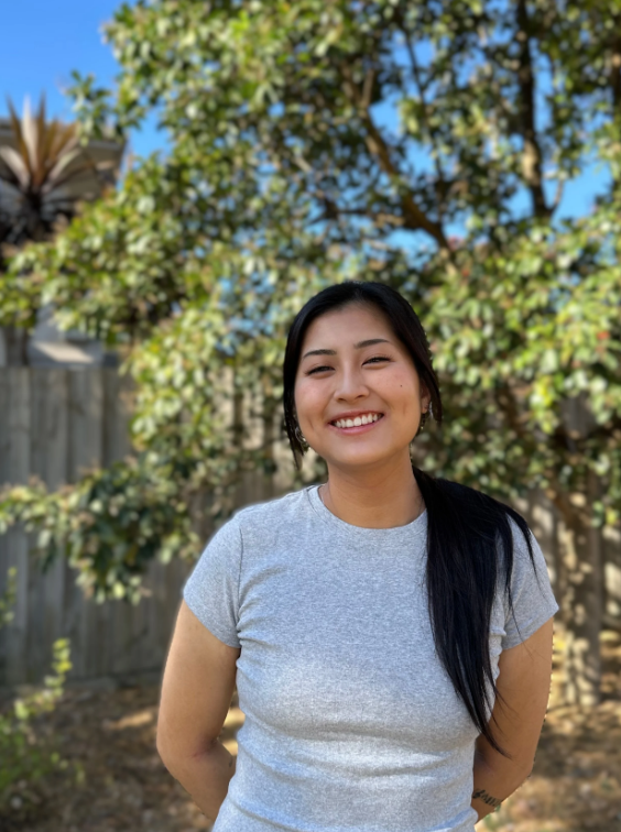 Smiling person standing outdoors in a sunny yard with trees and a wooden fence in the background.