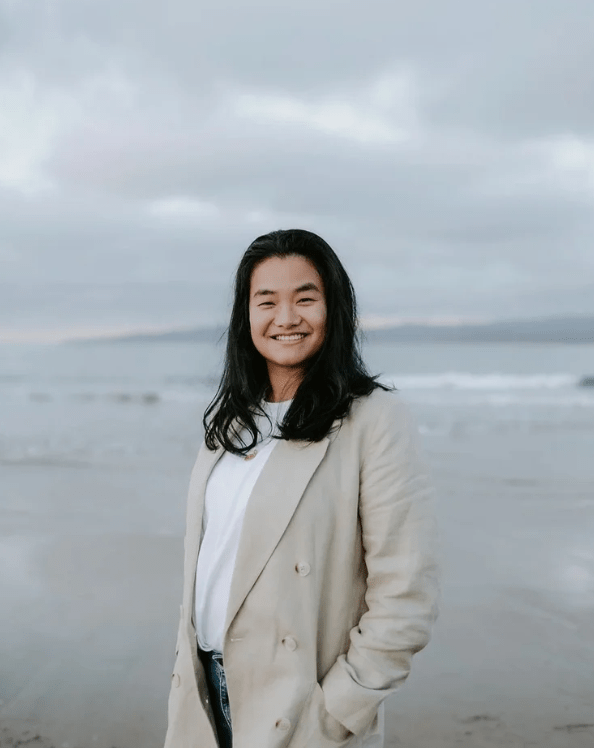 Smiling person with long dark hair in a beige blazer standing on a beach under a cloudy sky.