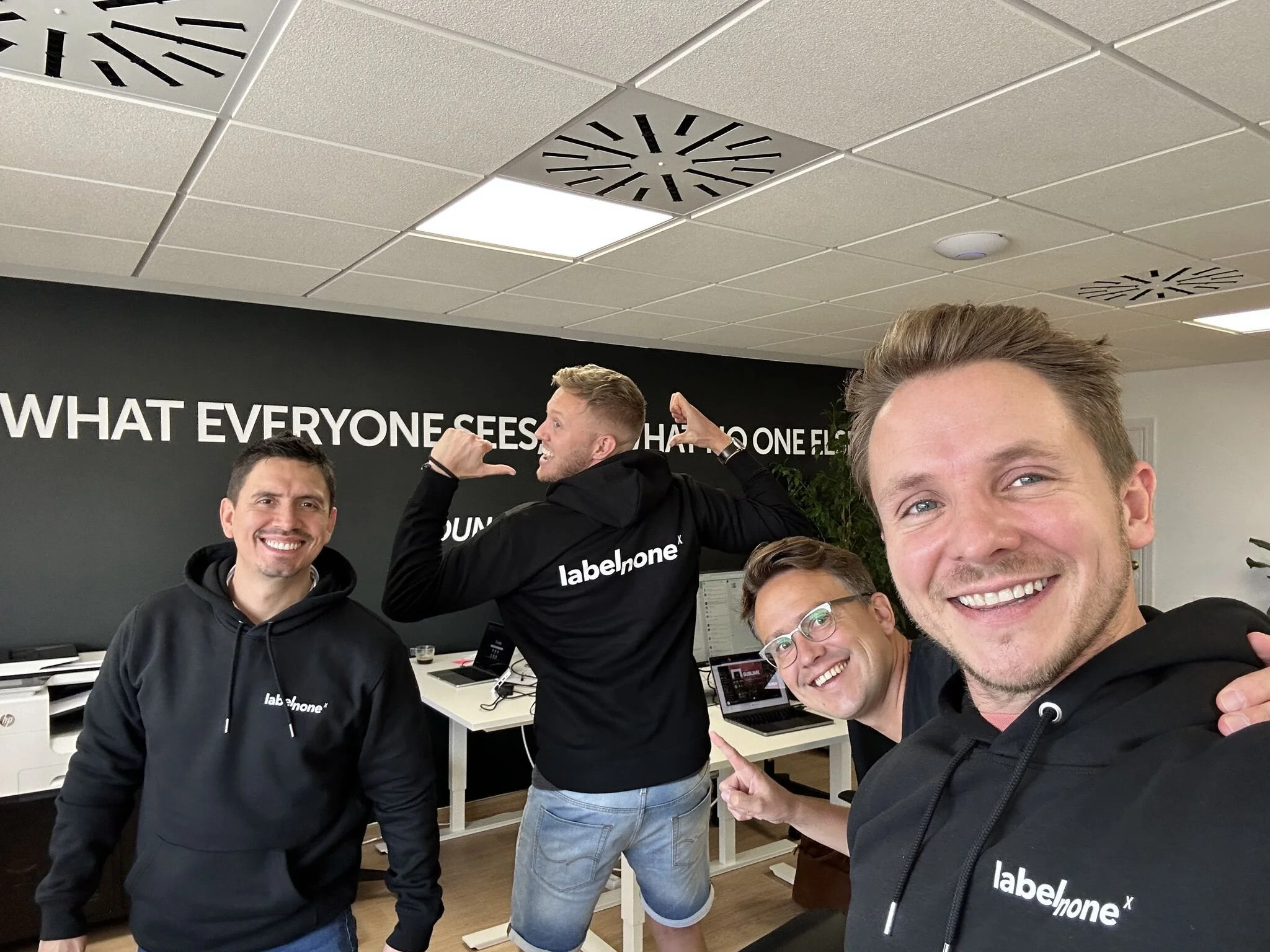 Four smiling men in a modern office. Two men are wearing black hoodies with 'labelnone' logos, one pointing to a wall, and another taking a selfie. The wall behind them has a large phrase, partially visible, reading 'WHAT EVERYONE SEES...'. There are desks with computers and office equipment in the background.