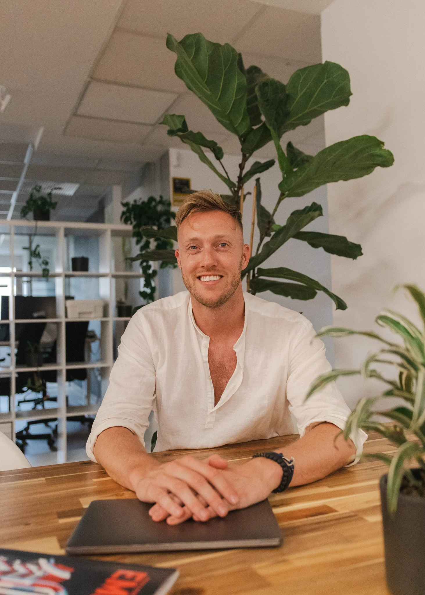 A smiling man in a white shirt sitting at a wooden table with a closed laptop, surrounded by green plants in an office setting.