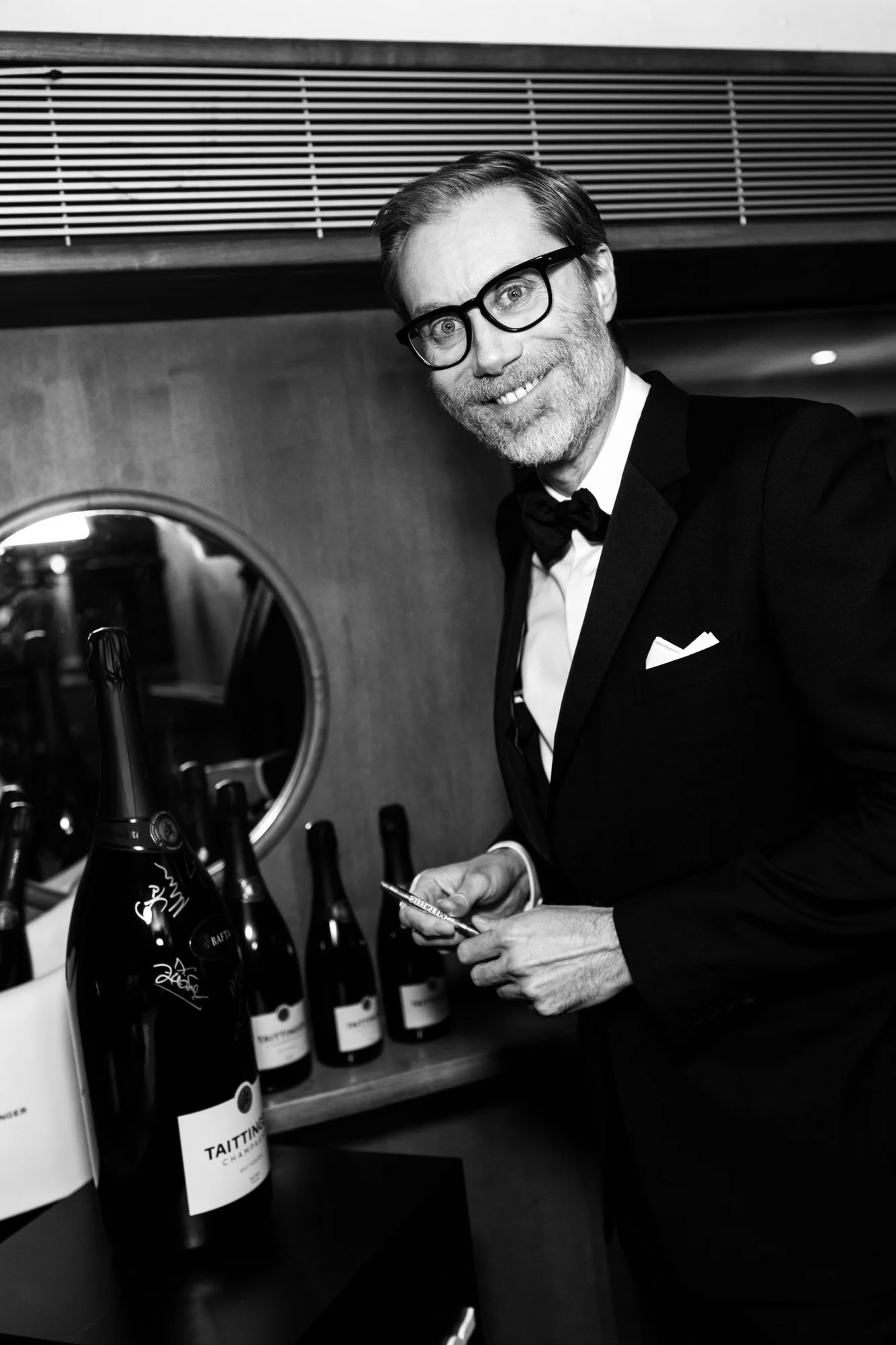 Stephen Merchant smiling backstage beside signed champagne bottles at the 2025 BAFTA Film Awards, photographed in black and white.