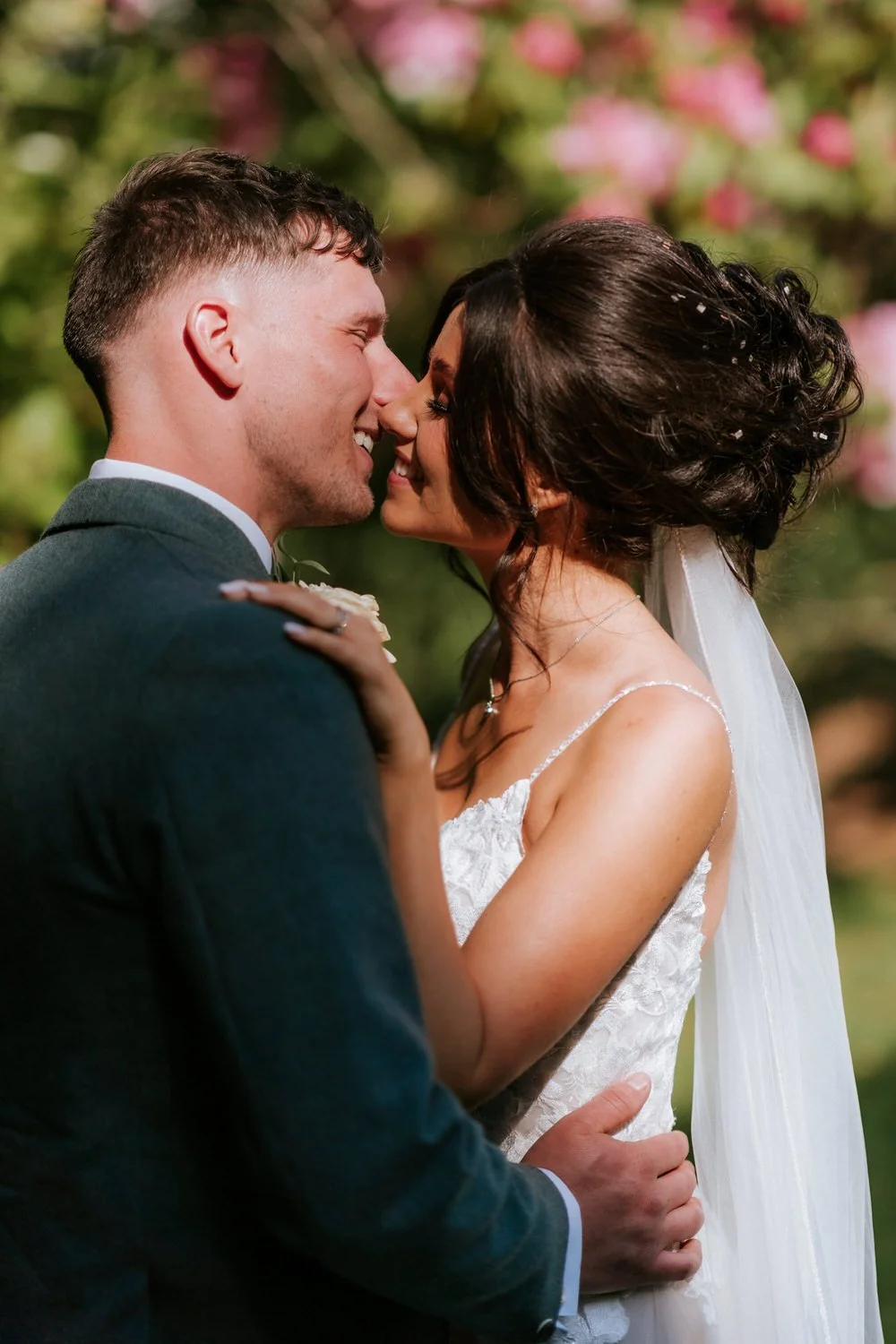 A newlywed couple about to kiss outdoors with a blurred green and pink floral background, the bride in a white wedding dress and veil, the groom in a dark gray suit.