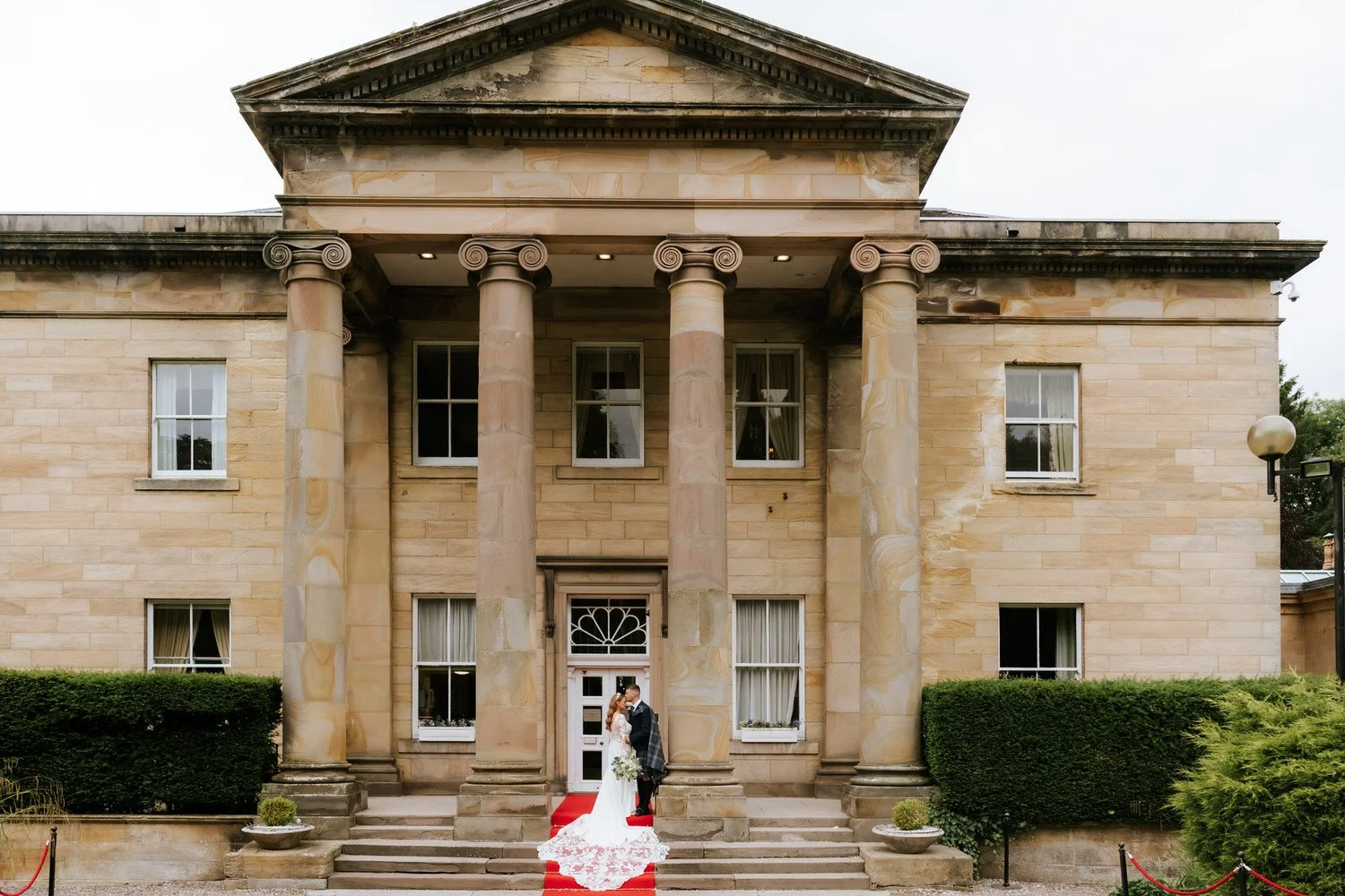 A bride and groom standing on a red carpet outside a large, historic stone building with tall columns and multiple windows, during a wedding.