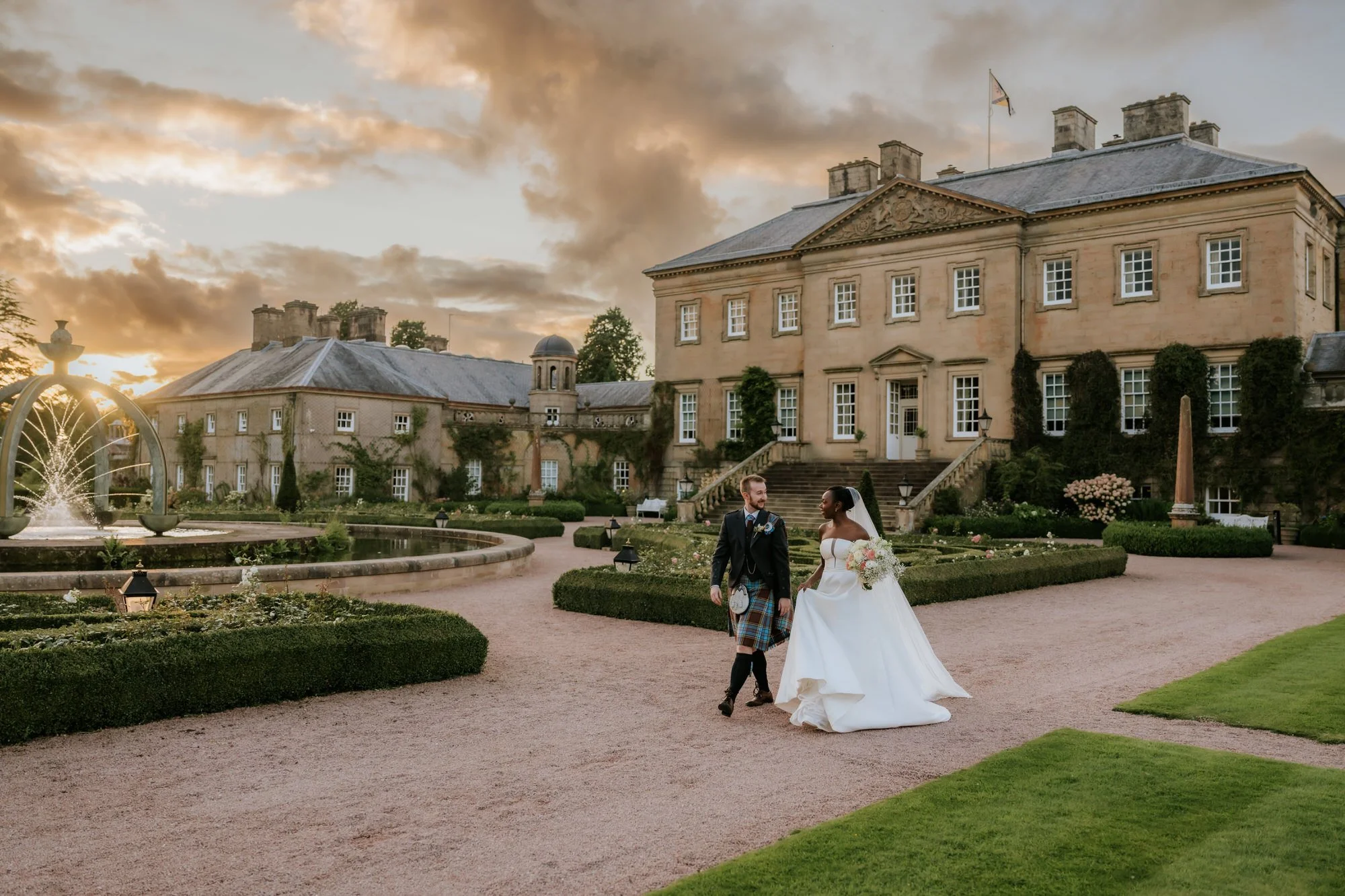 A newlywed couple walking in front of a historic mansion with a landscaped garden and fountain during sunset.