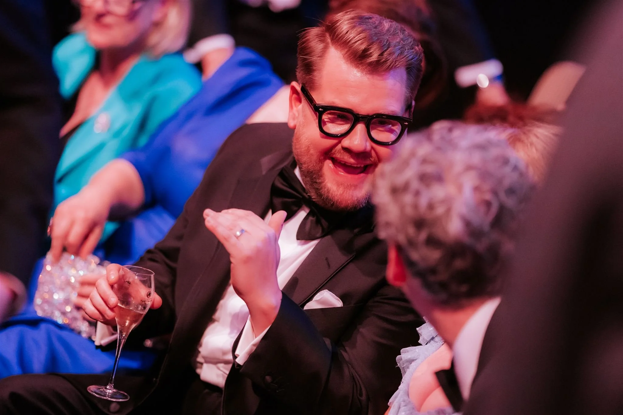 A man in a tuxedo with glasses holding a champagne glass, smiling and engaging in conversation with an older woman with gray hair at a formal event.