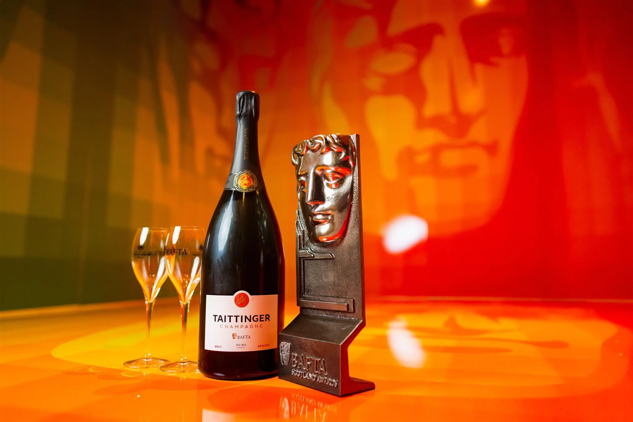 A bottle of Taittinger champagne with two glasses, and an award trophy with a face sculpture on a brightly lit orange surface against a backdrop with a similar face design.