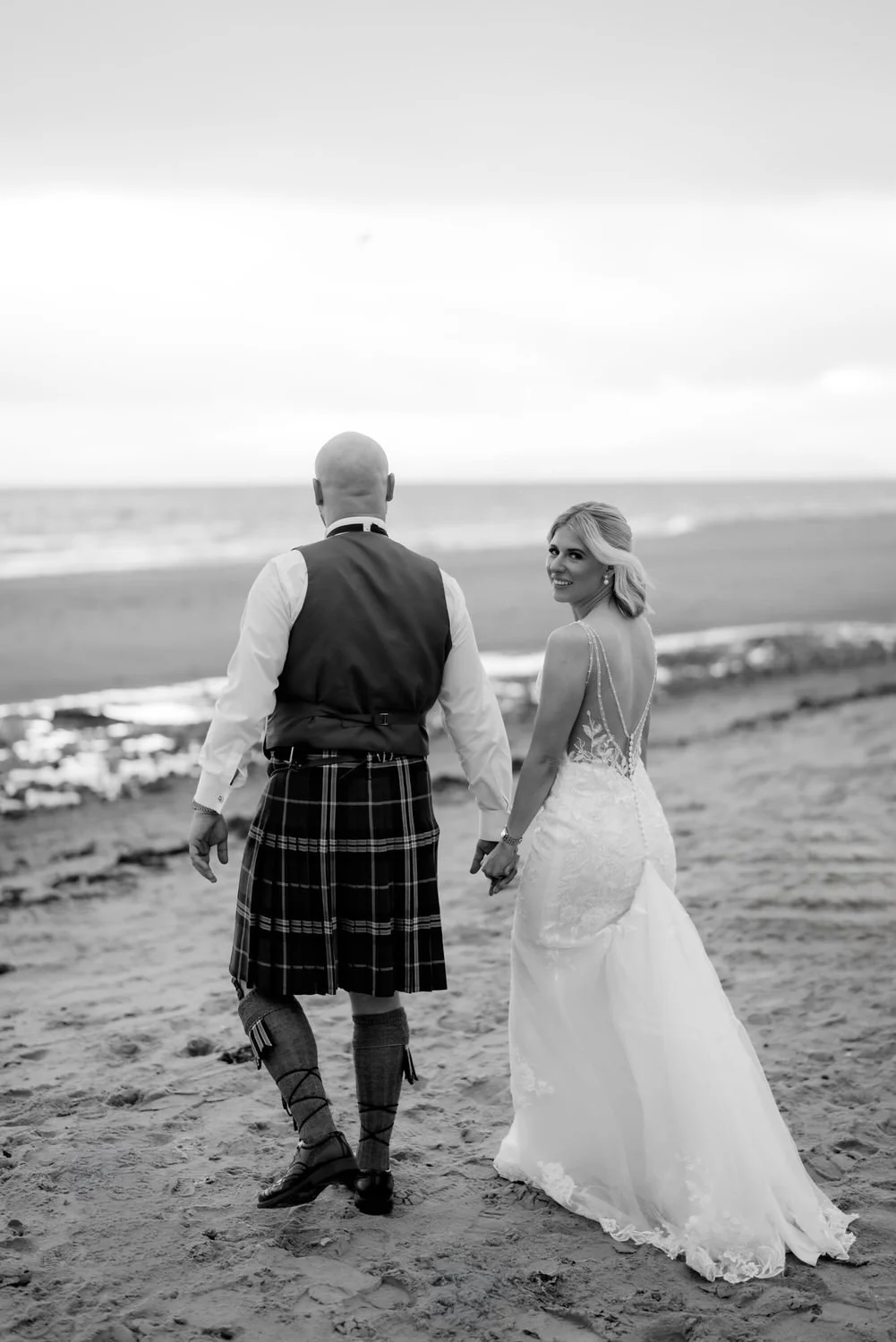 A bride and groom walking hand in hand on the beach, holding hands, with the bride smiling at the camera, in black and white.