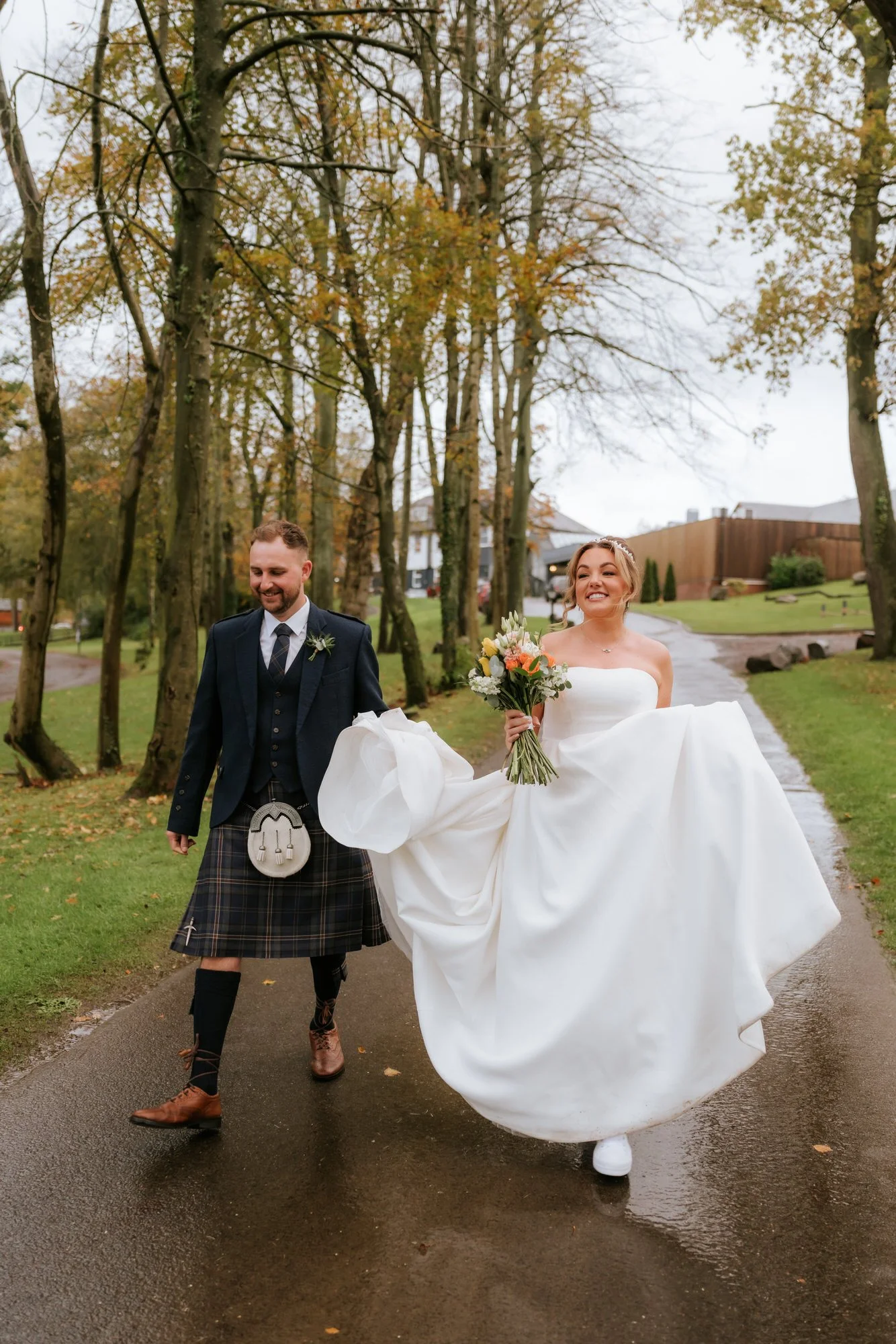 Bride in a white wedding gown holding a bouquet walking with a groom in a kilt and dark jacket on a wet pathway in a park with autumn trees.
