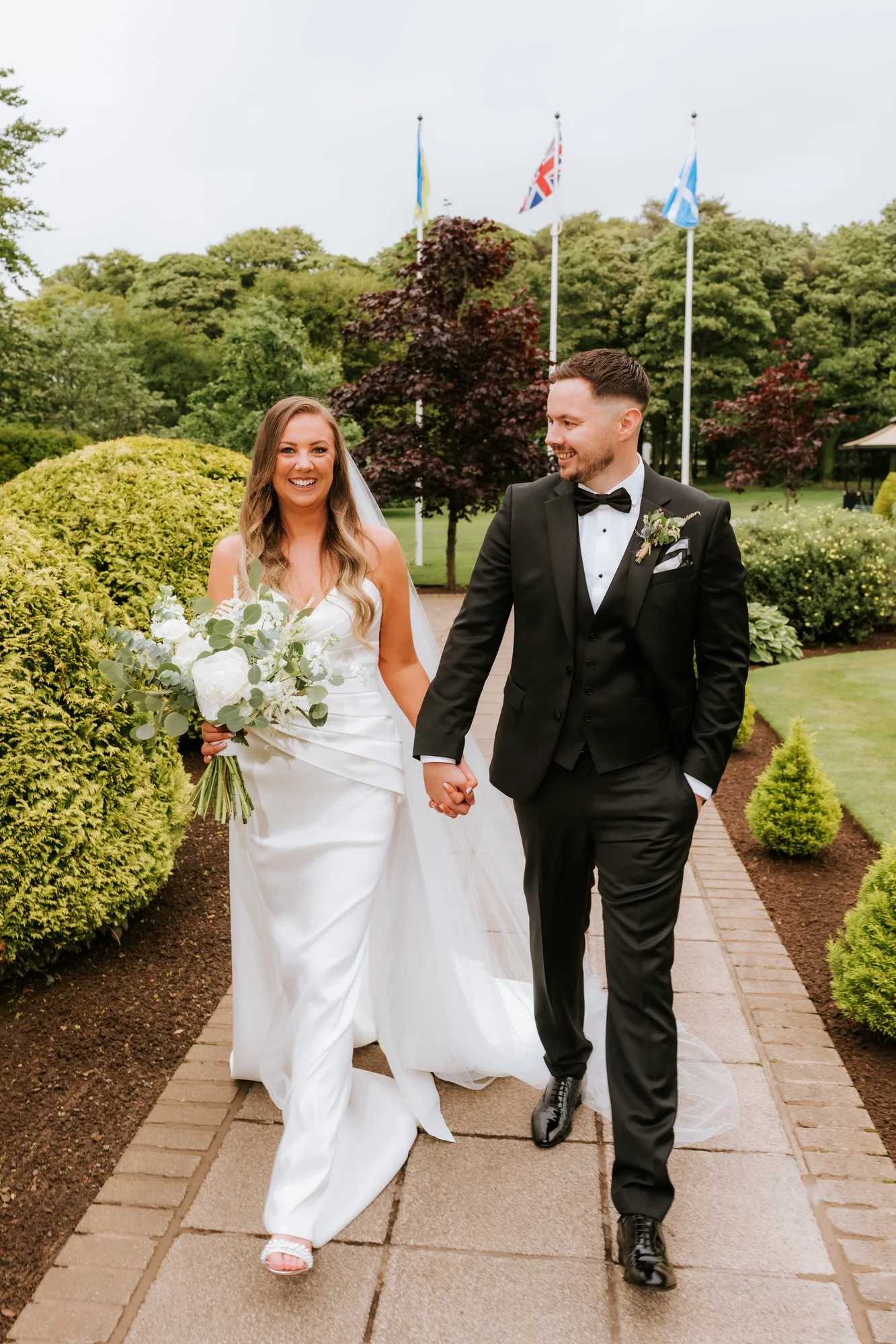 A newlywed couple walking hand in hand outdoors on a wedding day. The bride is in a white wedding gown holding a bouquet of white flowers and greenery; the groom is in a black tuxedo with a bow tie. They are smiling happily with flags flying in the b