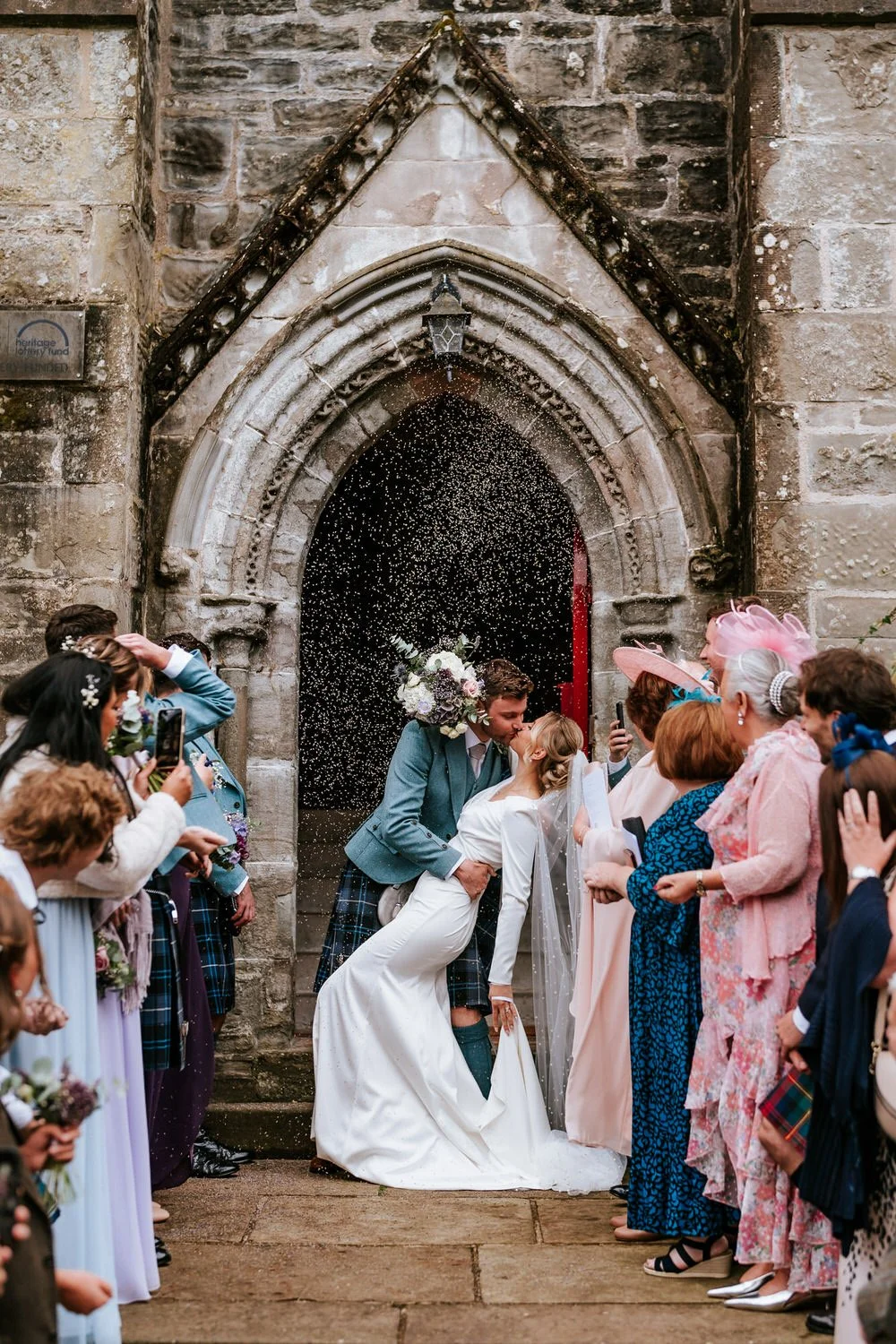 A bride and groom share a kiss outside a stone church during their wedding, with guests around them celebrating and taking photos, with confetti falling.