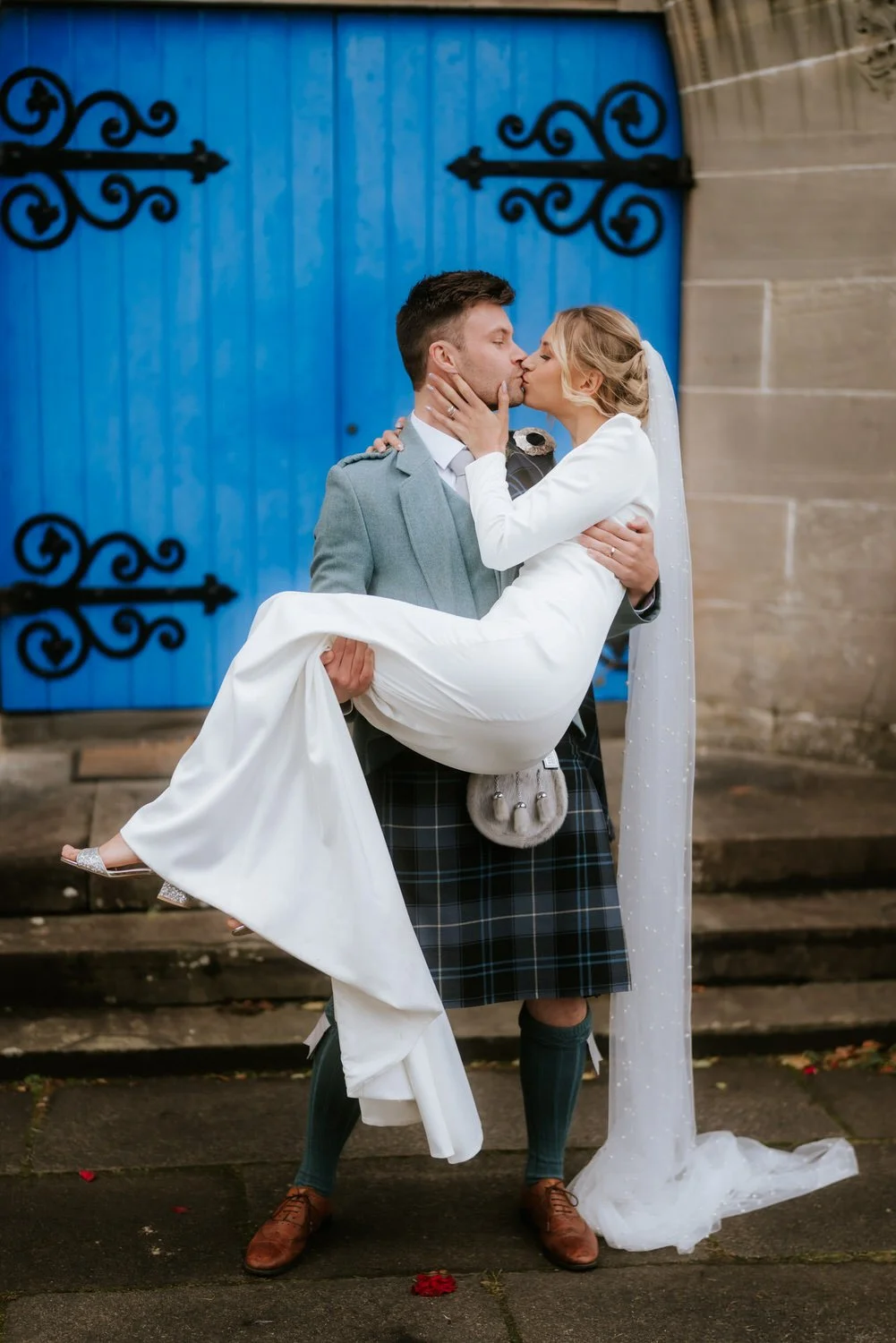 A newlywed couple sharing a kiss outdoors. The groom, dressed in a gray jacket and kilt, carries the bride in white wedding attire in his arms against a blue barn door.