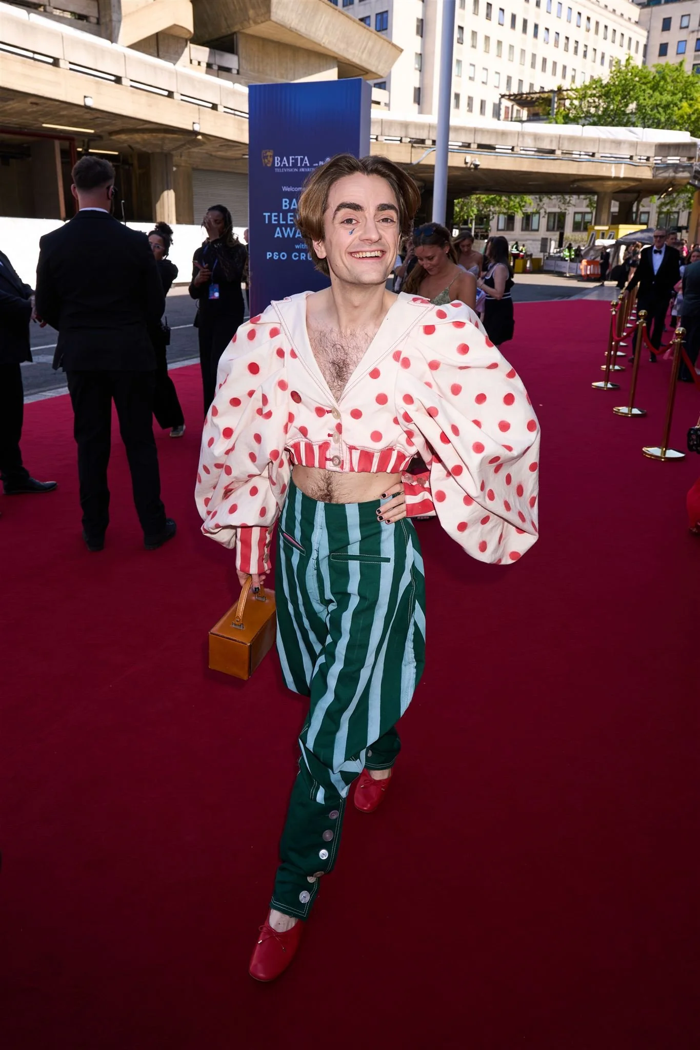 A person with brown hair is walking on a red carpet at an event, wearing a polka dot and striped outfit with red shoes, holding a small handbag, and smiling at the camera. There are other people and a sign in the background.