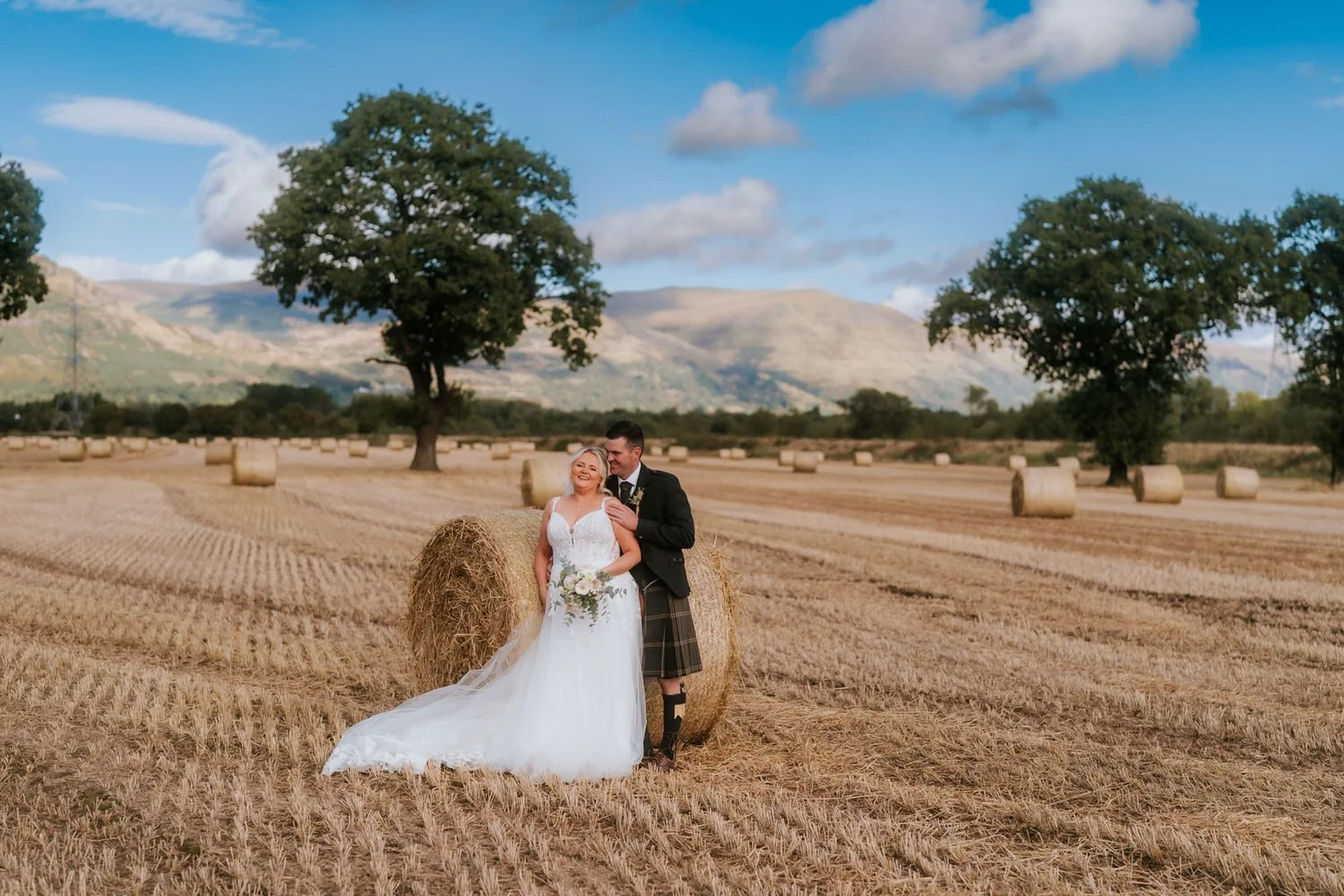 A bride and groom stand in a harvested field with hay bales, trees, and mountains in the background on a clear day.