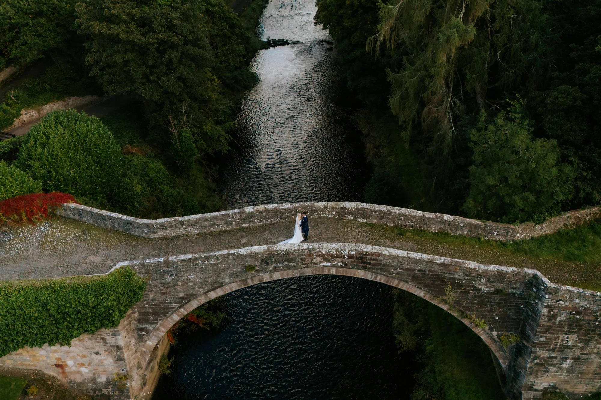 A bride and groom standing on a stone bridge over a river, surrounded by lush green trees.