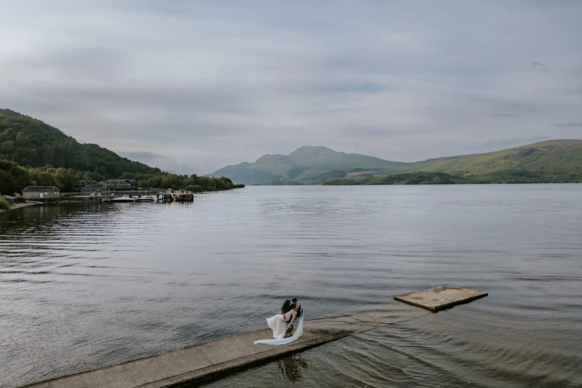 A bride and groom sharing a kiss on a dock by a lake with mountains in the background.
