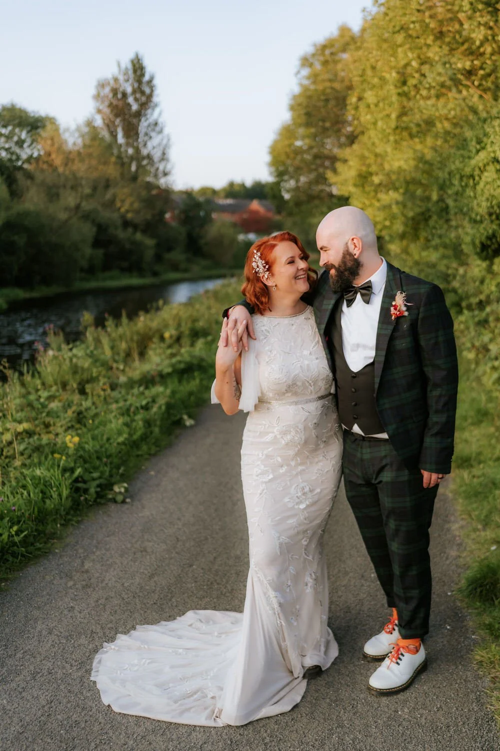 A bride and groom smiling and embracing outdoors on a pathway next to a river, with trees and greenery in the background.