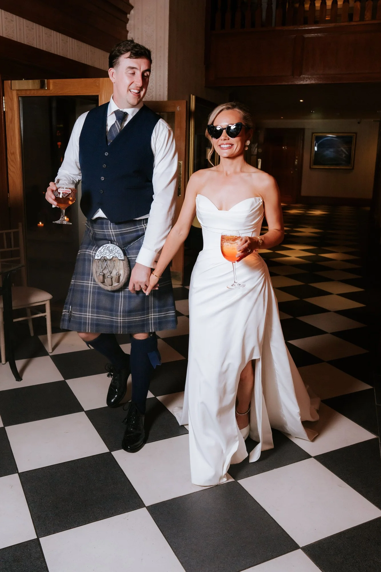 A woman in a wedding dress and sunglasses holding a drink, walking hand-in-hand with a man in traditional Scottish attire holding a drink, in a reception hall with checkered black and white floor.