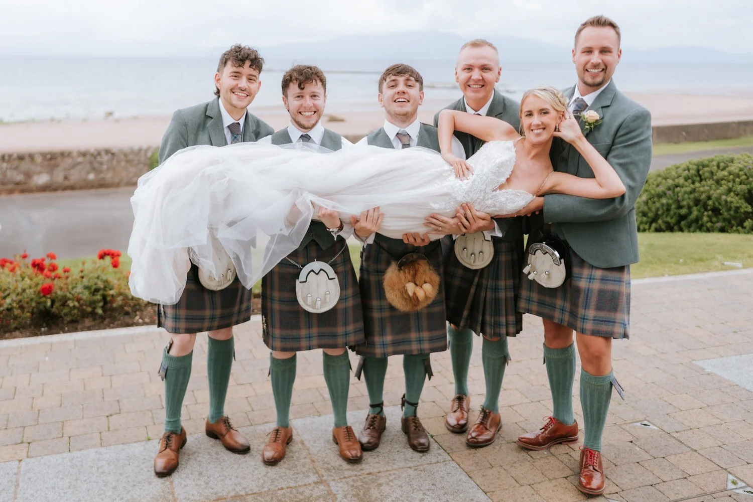 Group of six men in kilts holding a bride in a white wedding dress outdoors by the water.