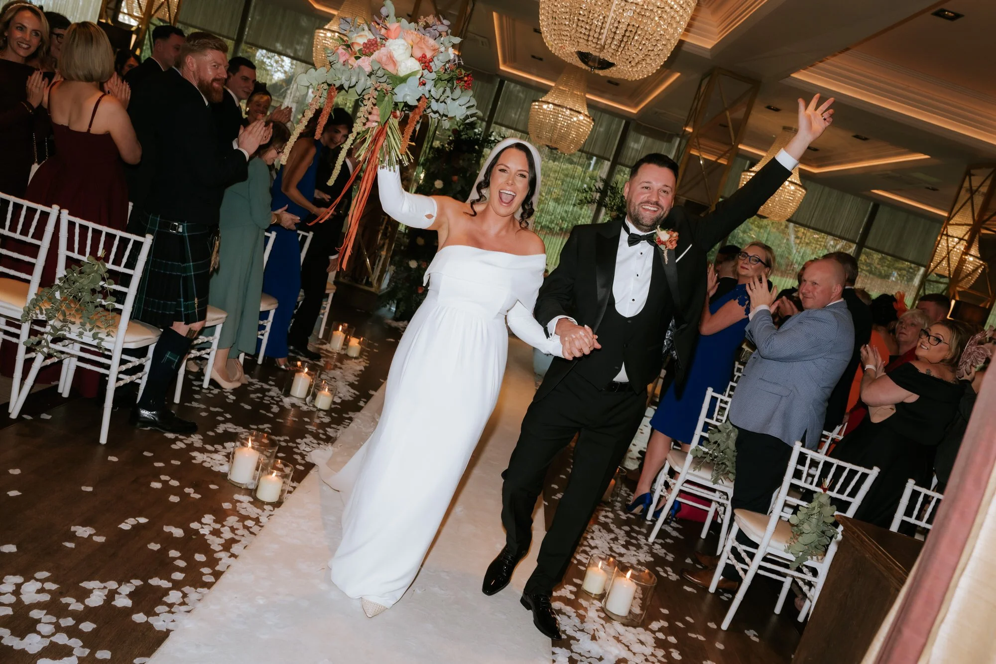 Happy bride in white dress and groom in tuxedo holding hands and smiling as they walk down the aisle at their wedding reception, surrounded by guests, candles, and flowers.