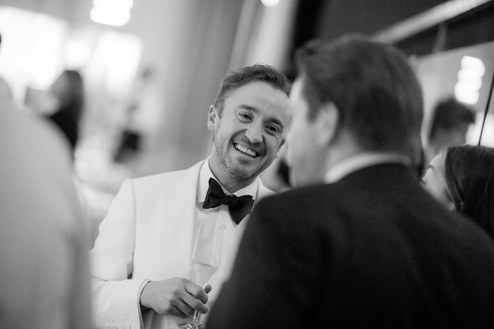 Tom Felton laughing with guests at the BAFTA dinner, photographed in black and white.