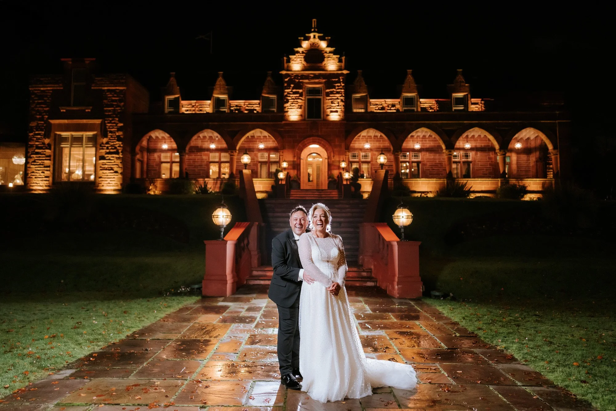 A newlywed couple in wedding attire, smiling and embracing, standing on a wet stone walkway in front of a large, illuminated historic mansion at night.