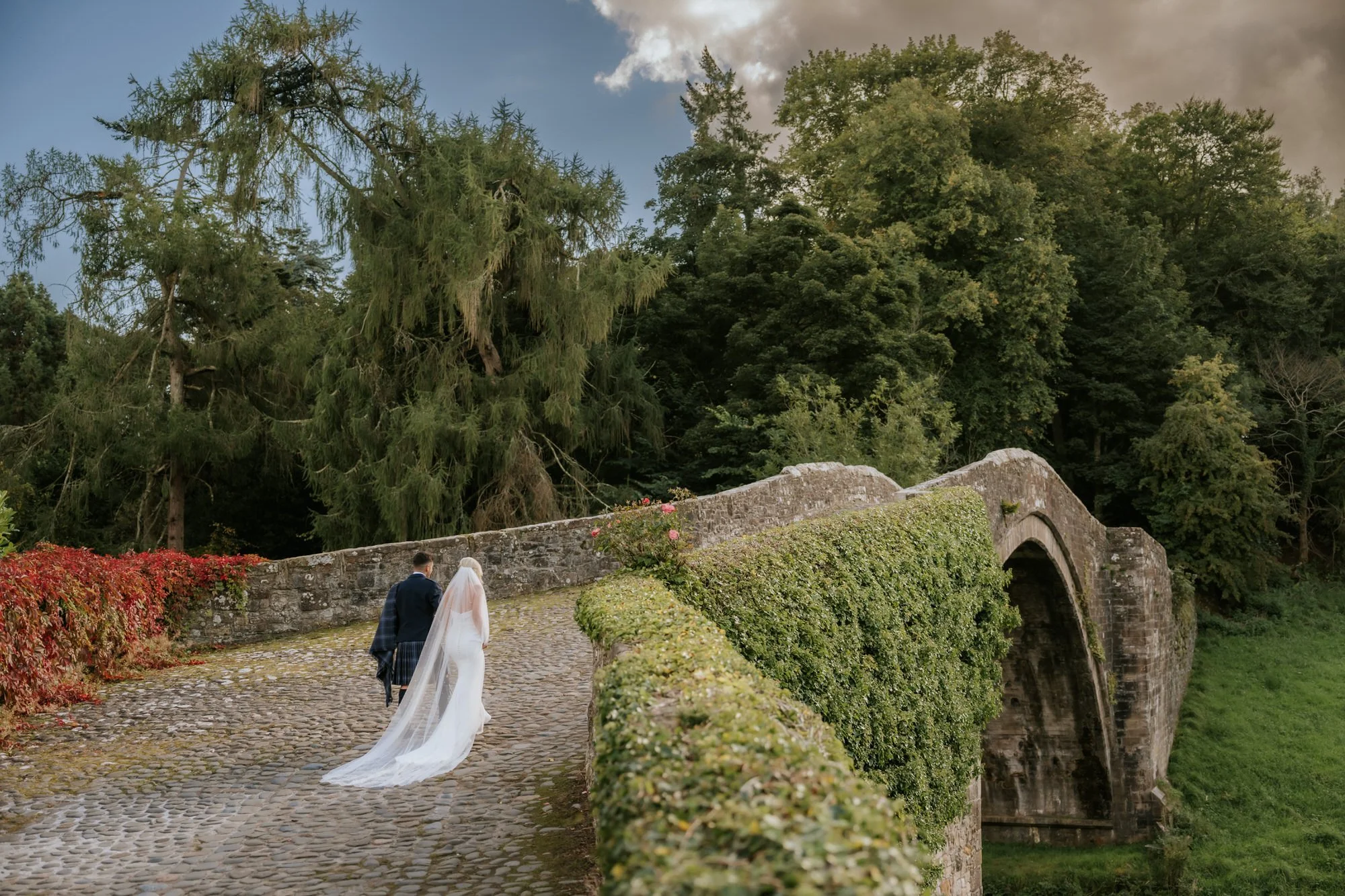 A bride and groom walking on a cobblestone path over a stone bridge, surrounded by green trees and climbing ivy, during late afternoon or early evening.
