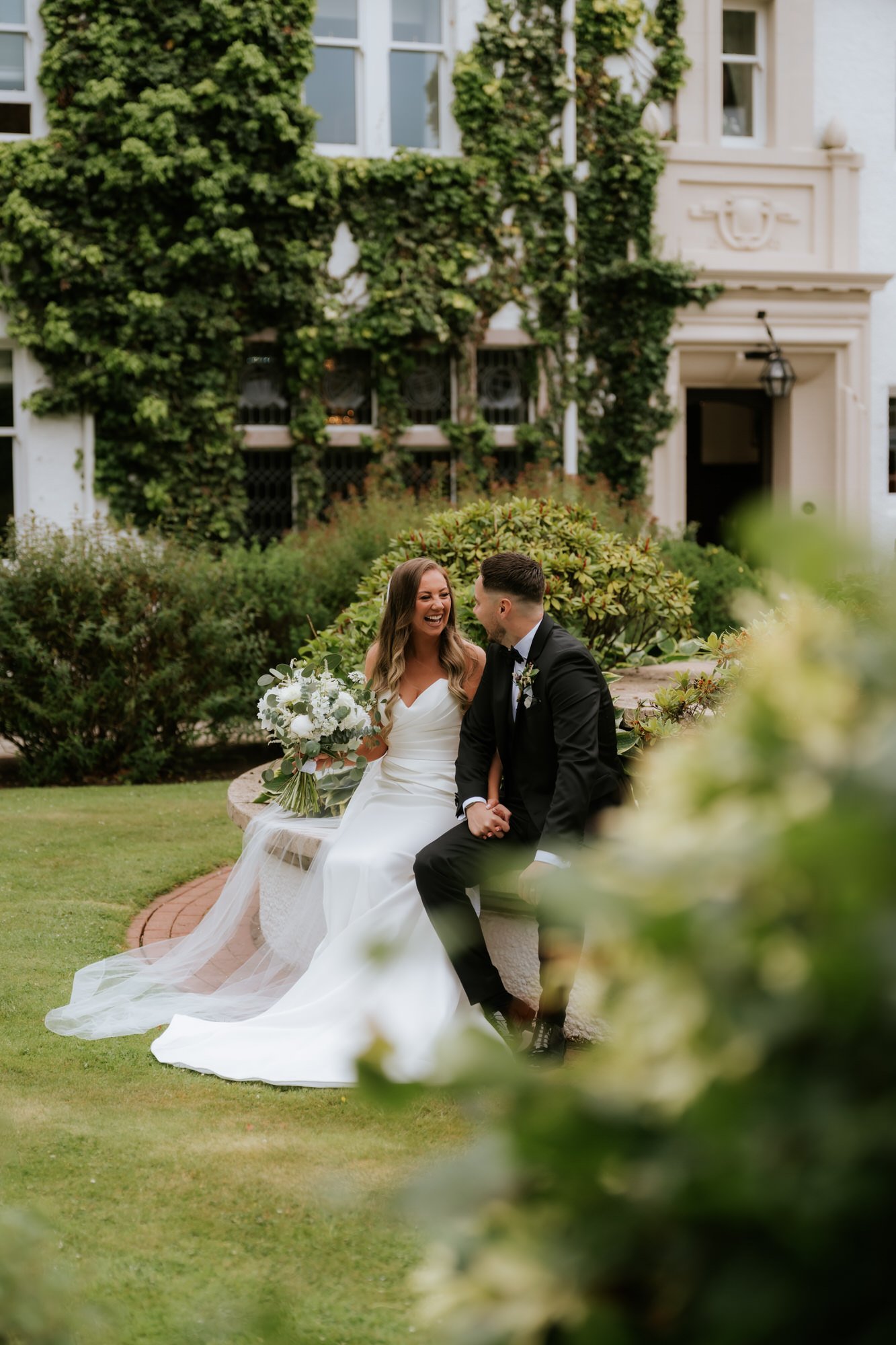 A newlywed couple sitting on a stone bench outdoors, holding hands and smiling. The bride is in a white wedding dress holding a bouquet, and the groom is in a black suit. Greenery and a white building are in the background.