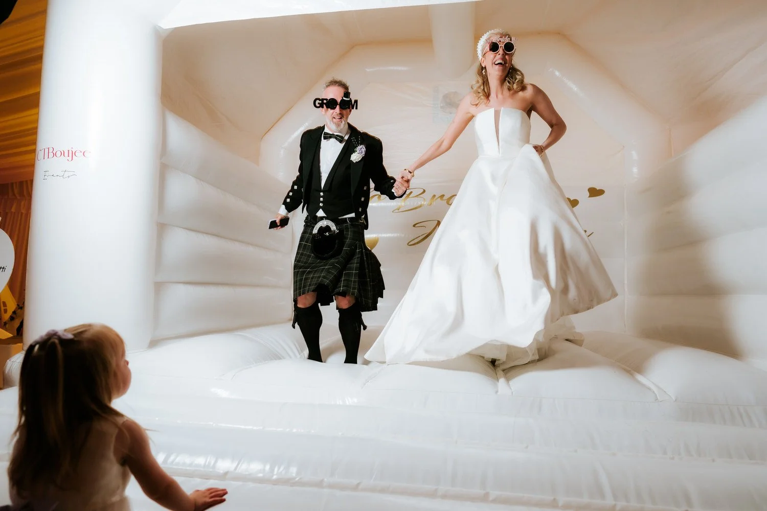A bride and groom on an inflatable wedding cake, holding hands and smiling, with a young girl watching from below.
