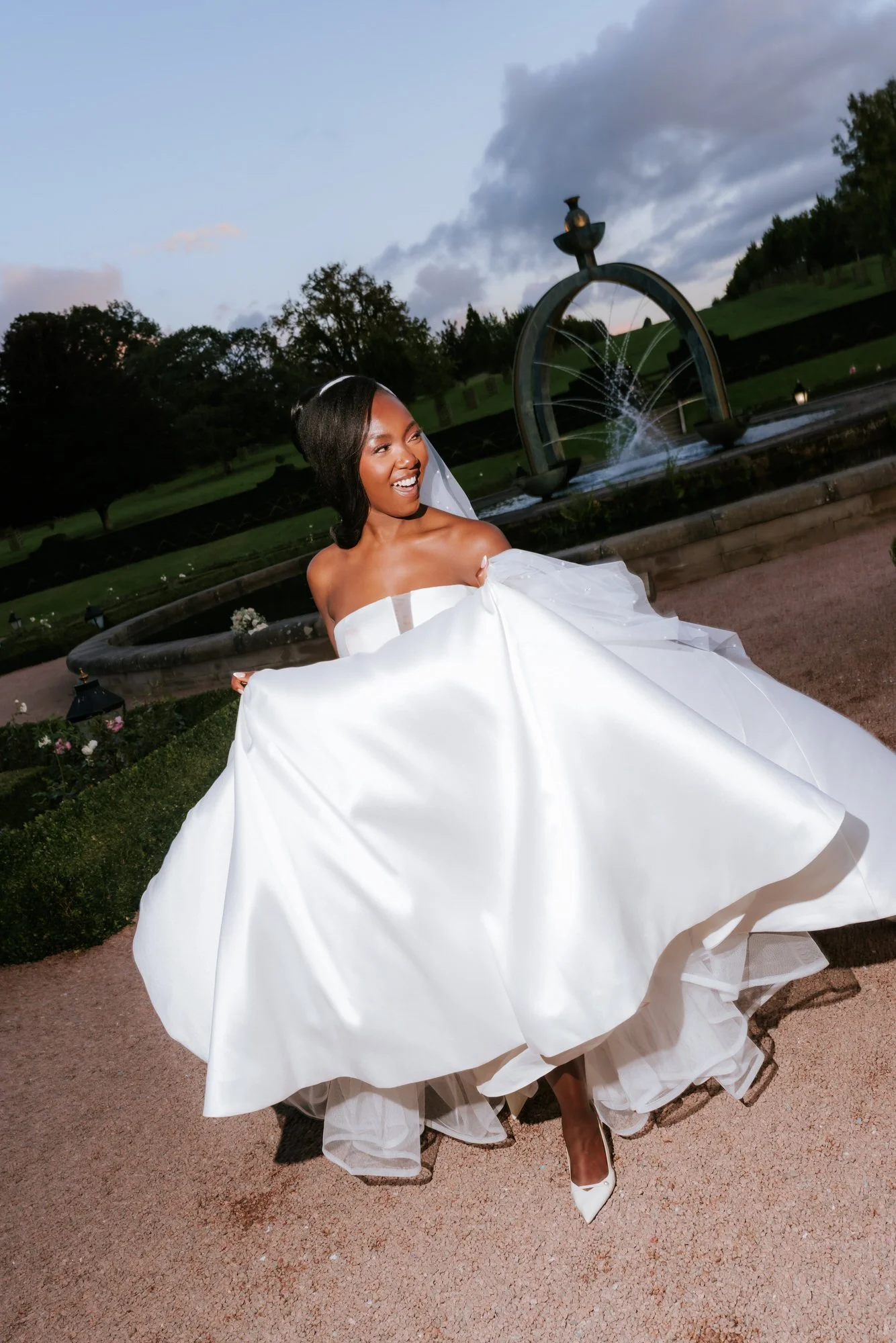 A bride in a white wedding dress smiling outdoors near a fountain at dusk.