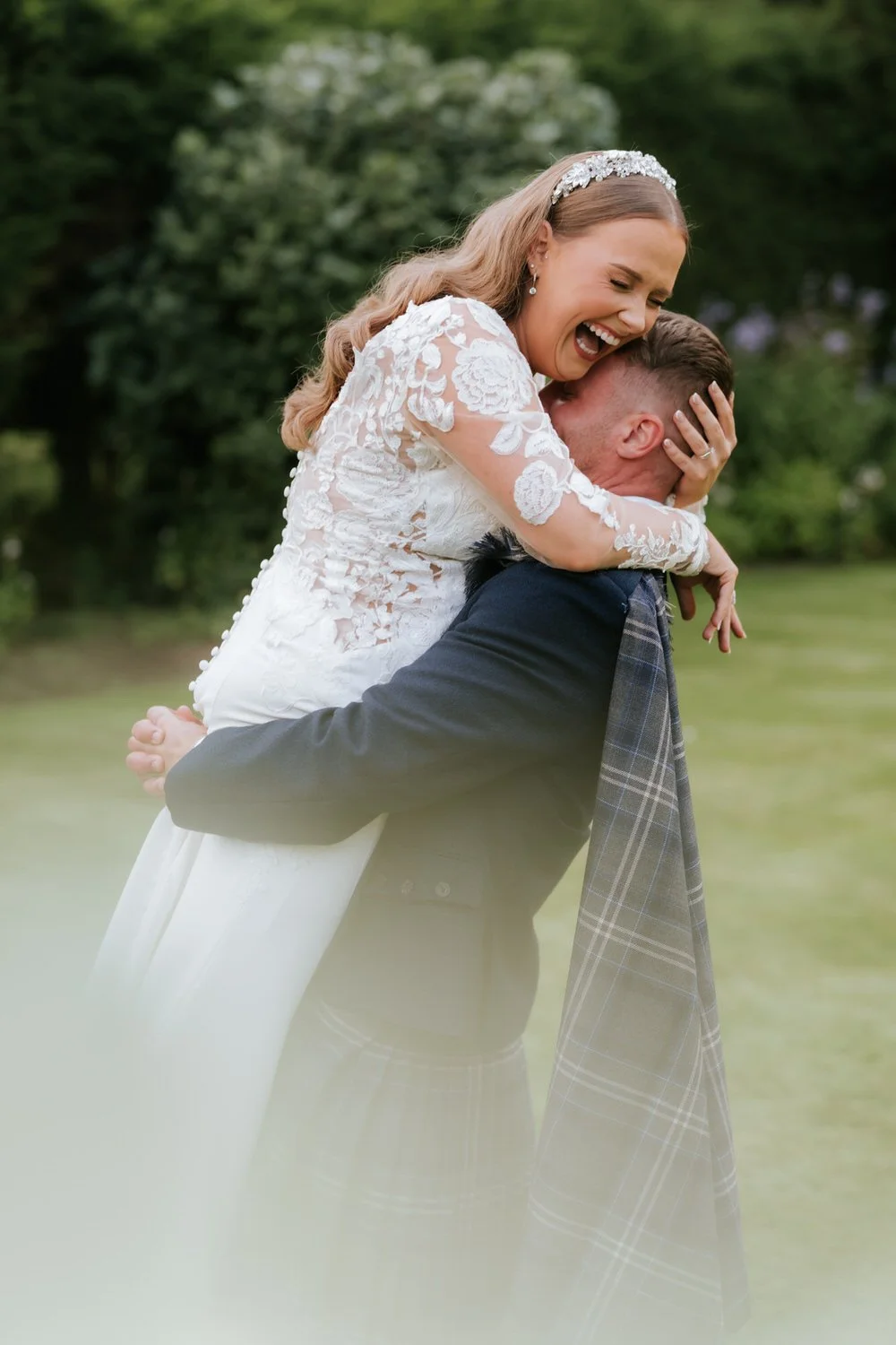 A bride with long blonde hair in a white lace wedding dress and a tiara, smiling and hugging a man outdoors in a green park, who is wearing a dark suit and a tartan kilt.
