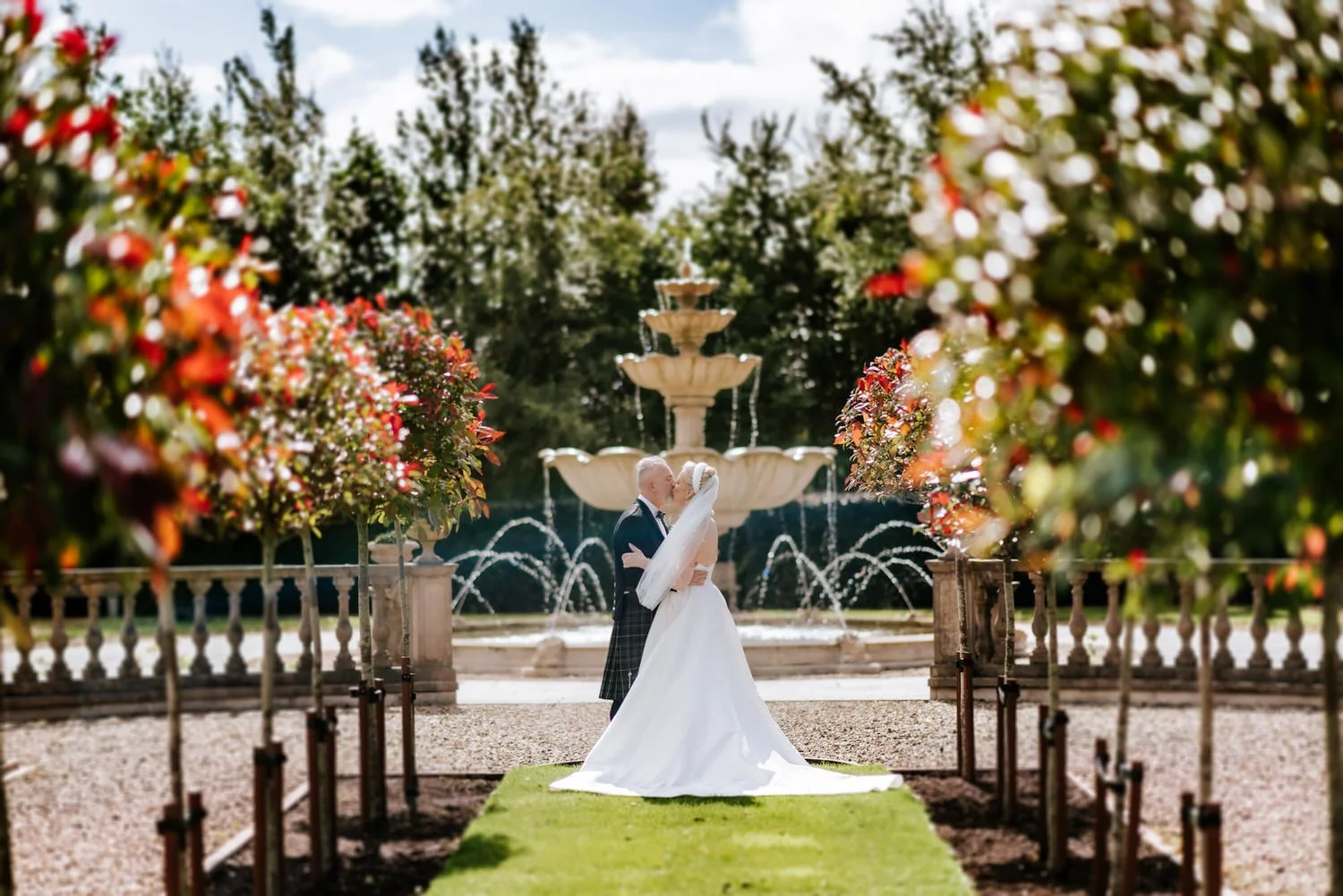 A bride and groom sharing a kiss in front of a fountain with trees and a fountain in the background, surrounded by blooming flowers.
