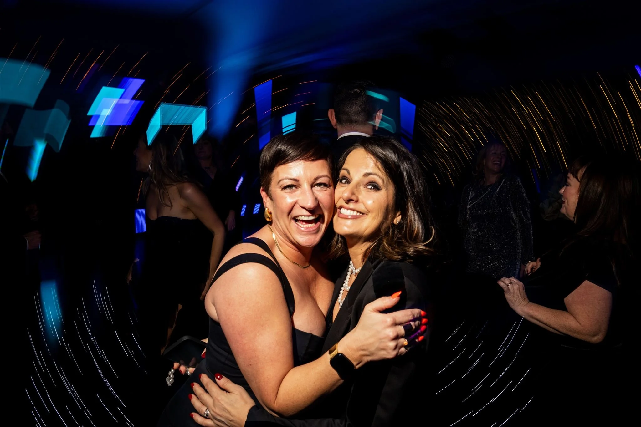 Two women in black dresses smiling and hugging at a lively party with colorful lights and blurred background of other guests.