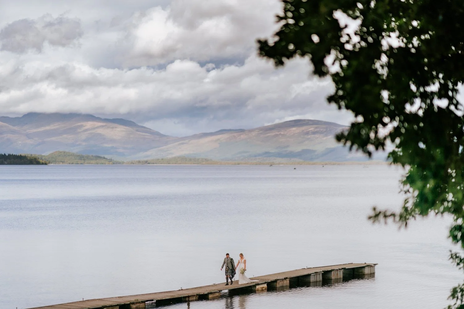 A bride and groom walking hand in hand on a dock over a peaceful lake with mountains and a cloudy sky in the background, partially framed by a tree on the right side.