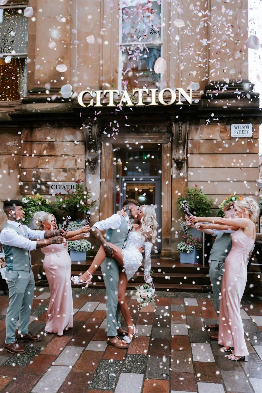 Bride and groom kissing in front of a building marked 'CITATION' while being showered with confetti by wedding guests holding party poppers, with flowering plants and decorative elements around.