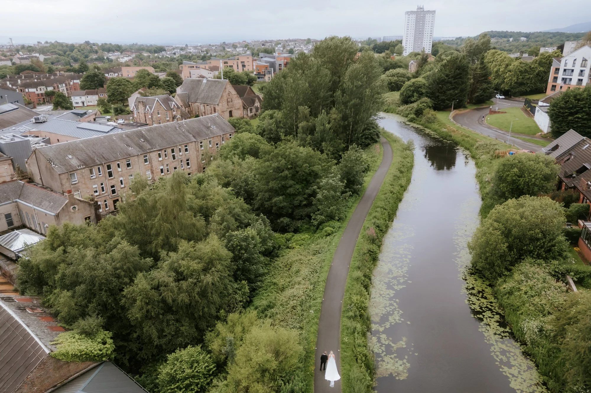 A couple walking on a narrow path beside a river in a green city park, with residential buildings and trees all around.