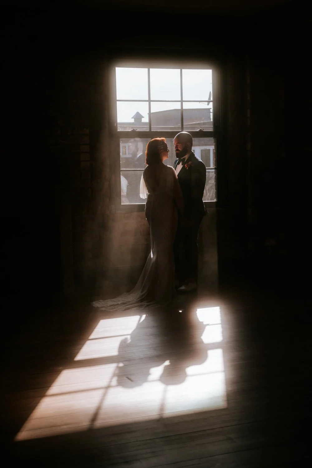 A bride and groom standing close together in front of a large window, backlit by sunlight, creating a silhouette effect with their faces nearly touching.