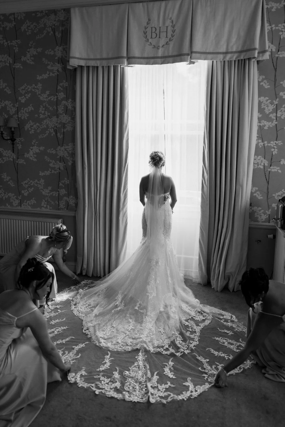 A bride in a wedding dress stands by a window, surrounded by four women helping her with her dress. The room has floral wallpaper, curtains, and soft lighting.