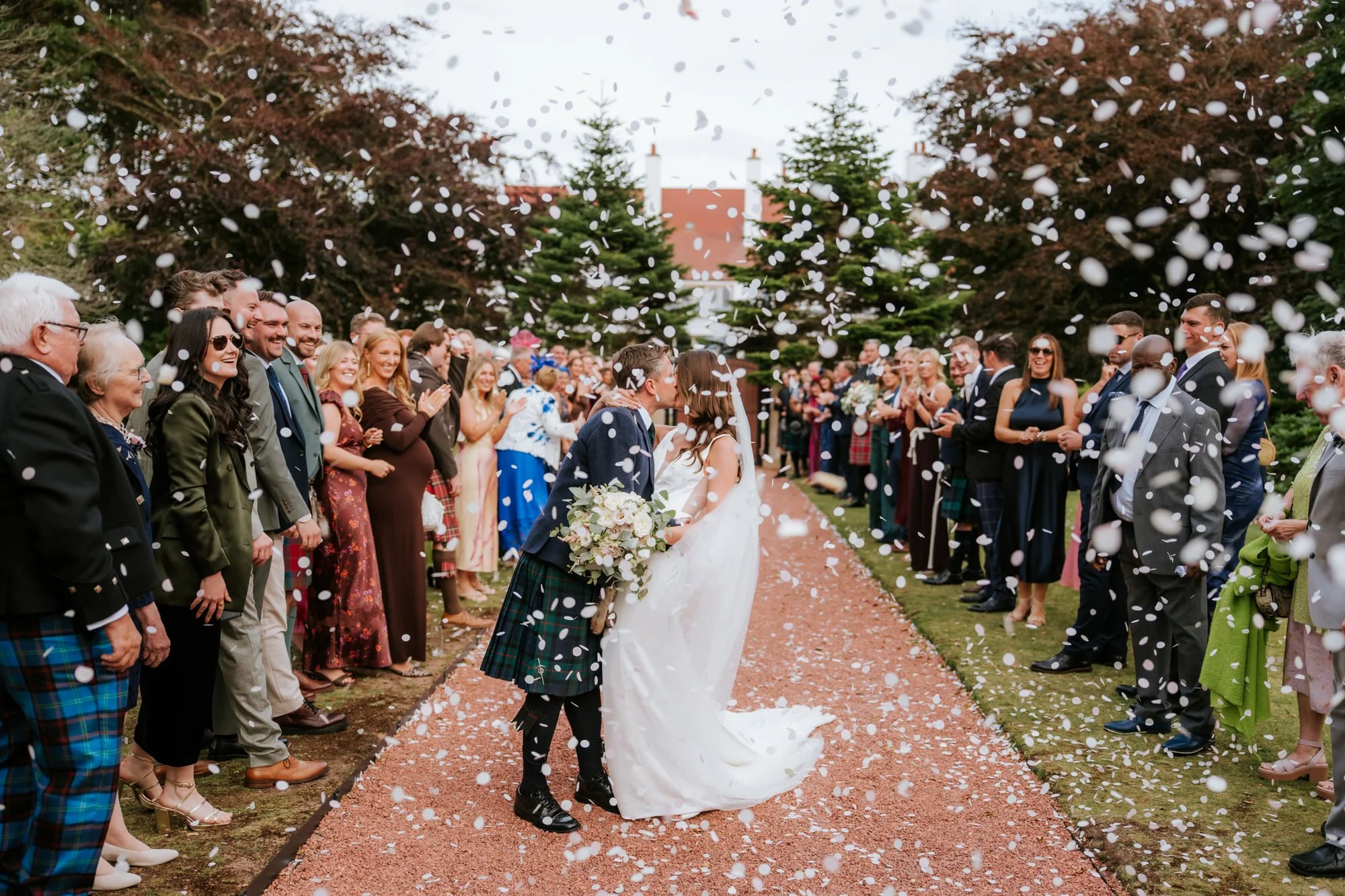 A bride and groom kiss while guests celebrate and throw confetti at an outdoor wedding ceremony.